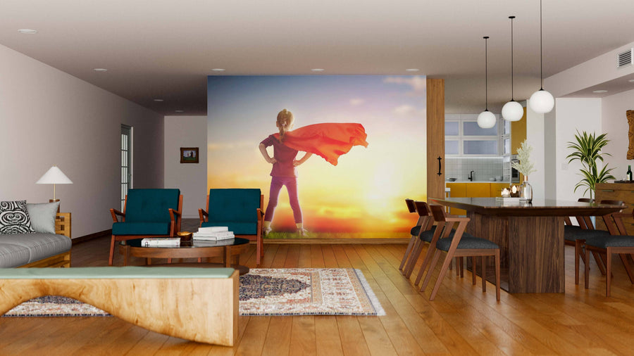 A modern living/dining room with wood floors, stylish chairs, and pendant lights is brightened by the Girl Plays Superhero Wall Mural, showcasing a child in a superhero pose against a sunset sky for a touch of warmth and whimsy.