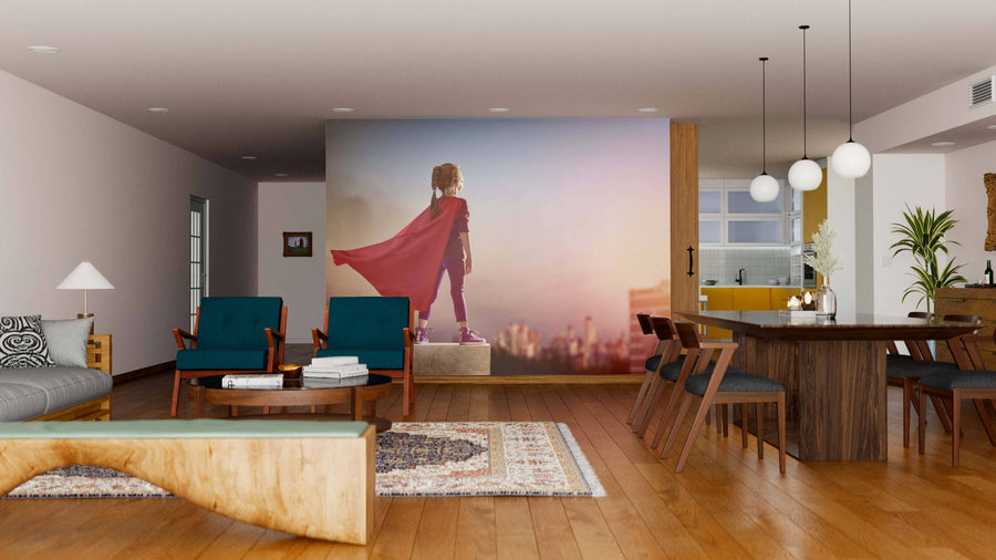 A modern living-dining room with wood floors, teal chairs, a rug, and wooden dining table features the Girl Superhero Wall Mural, showing a child in a red cape gazing at a pastel city skyline. The kitchen is visible in the background.
