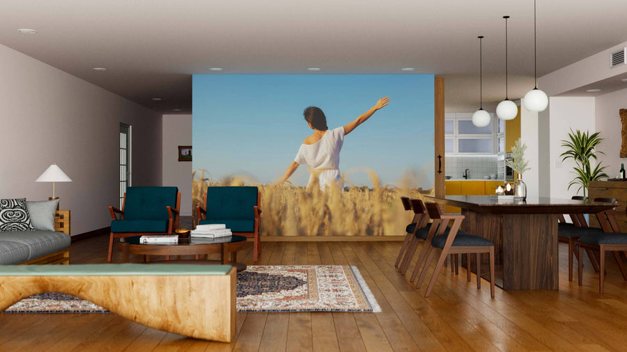 A modern open-concept living and dining room with wood floors and mid-century furniture features the Girl in Wheat Field Wall Mural, showing a woman in a white dress standing in wheat under a clear blue sky, one arm raised.