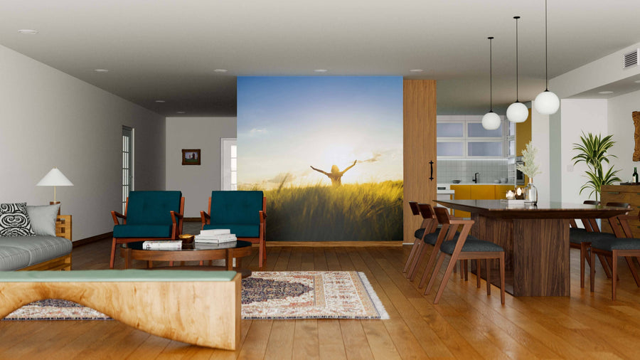 A modern open-plan living and dining room with wood floors, teal chairs, a wooden table, rugs, and pendant lights features the Girl on Sunset Wall Mural as a focal point, depicting a person in a sunlit field, with a yellow kitchen in the background.