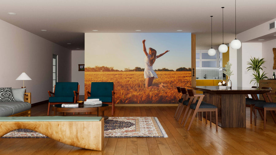 A modern living and dining area with wooden floors, teal chairs, and a gray sofa is enhanced by the Golden Field Bliss Wall Mural, featuring a woman in a white dress dancing in a golden field, alongside sleek lamps and a wooden dining table.