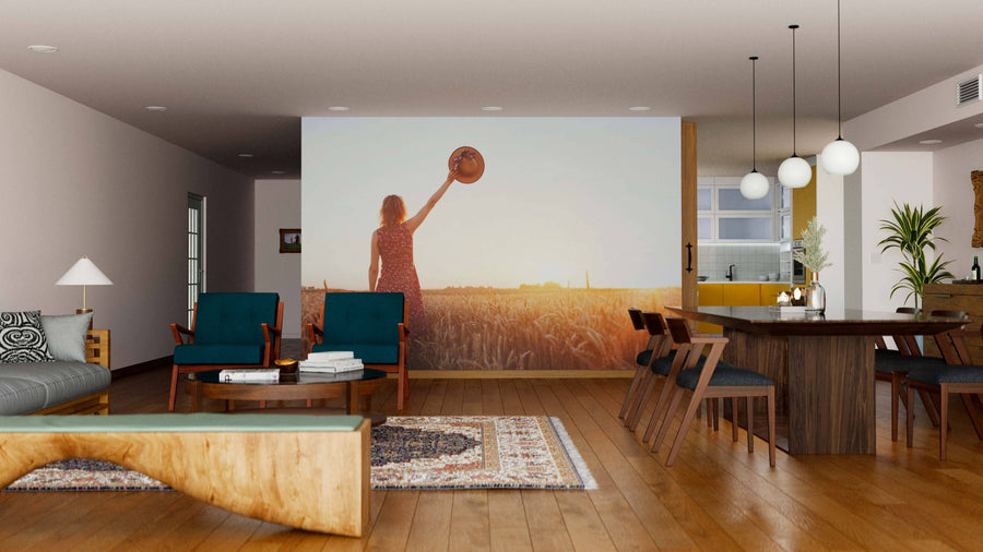 A modern living-dining room features wood floors, teal armchairs, a wood coffee table, and the Golden Fields Wall Mural depicting a person in a field at sunset. Pendant lights illuminate the dining table with stylish chairs.