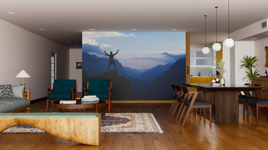 A modern living and dining room with wooden floors, stylish furniture, and the Man Hiking Wall Mural depicting a triumphant figure on a mountain peak. Warm lighting and cozy decor complete the space against misty blue valleys.