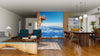 A modern living-dining room with wooden floors and mid-century furniture features the Man on the Cliff Wall Mural, depicting a climber above clouds and mountains to create an illusion of open views. The kitchen is visible in the background.