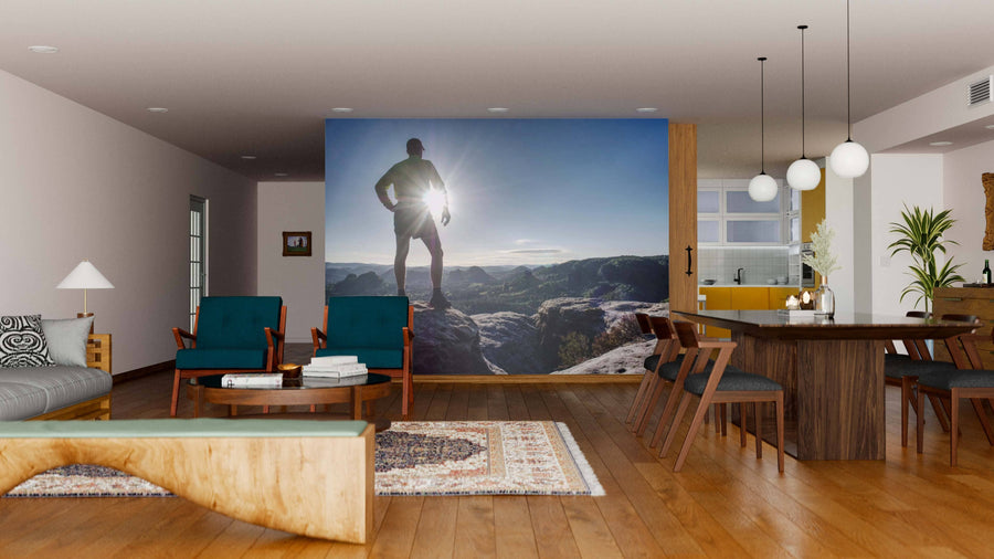 A modern living-dining room features wood floors, mid-century furniture, and an "Own the Summit Wall Mural" showing a hiker on a mountain at sunrise. Sunlight pours in from windows while globe pendant lights illuminate the dining area.