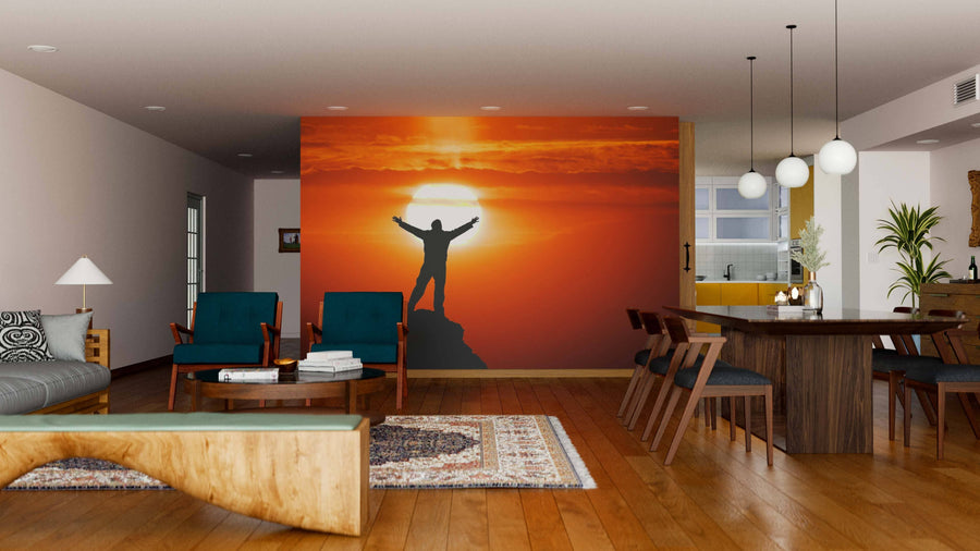 A modern living-dining room with wooden floors, dark blue chairs, and a large Peak of Mountain Wall Mural featuring a person standing triumphantly on a mountain at sunset with orange clouds and a glowing sun.