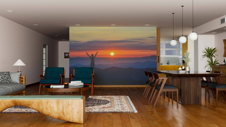 A modern living-dining room with wood furniture, a patterned rug, and pendant lights displays the Rise and Conquer Wall Mural—a peel and stick mural featuring a silhouetted person with raised arms against a vibrant mountain sunrise.