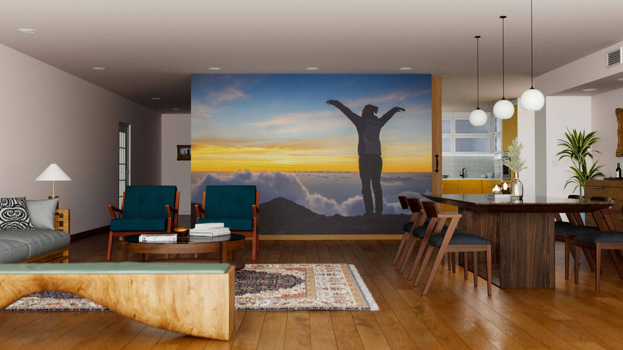 A modern living and dining area with wooden floors, teal armchairs, a wooden table, and contemporary lighting features the Sea of Clouds Wall Mural depicting a person with outstretched arms at sunrise above colorful mountain clouds.