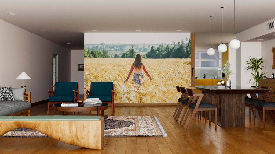 A modern living-dining room with wooden floors, teal armchairs, and a dining table features the Serene Countryside Wall Mural, depicting a woman walking through a golden wheat field under a blue sky for a natural, tranquil vibe.