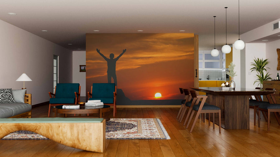Modern living-dining room with wooden floors and furniture, featuring the “Silhouette of Man at Sunset Wall Mural” on one wall. Green chairs, a wooden table, pendant lights, and a rug add warmth and style to the space.