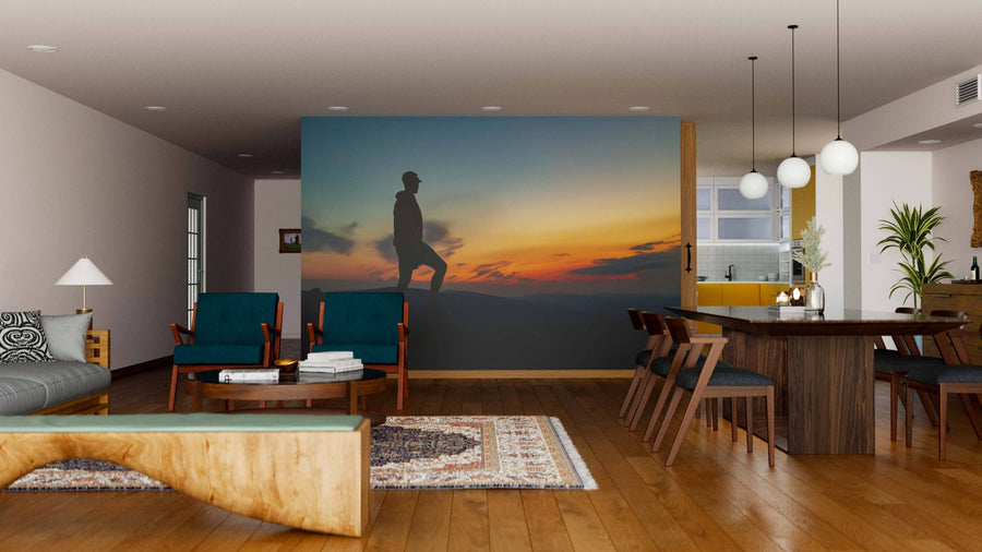 A modern living-dining room features wooden floors, dark teal chairs, and a wooden dining set. The Standing Above the Clouds Wall Mural on the room divider depicts a person on a hill at sunset, creating a focal point beneath round pendant lights.