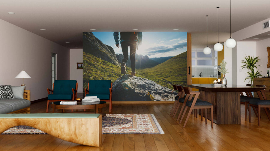 A modern living-dining room with wooden floors, green and teal furniture, and pendant lights features the striking Steps to Summit Wall Mural, showing a close-up of a hiker’s legs atop a rock in a sunlit mountain landscape.