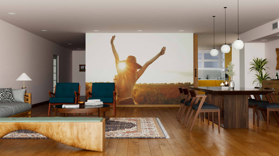 A modern, open-plan living and dining room features wooden floors, contemporary furniture, teal chairs, a wood dining table, pendant lights, large windows, and the Sunlit Joy Wall Mural depicting a person with arms raised in a sunlit field.