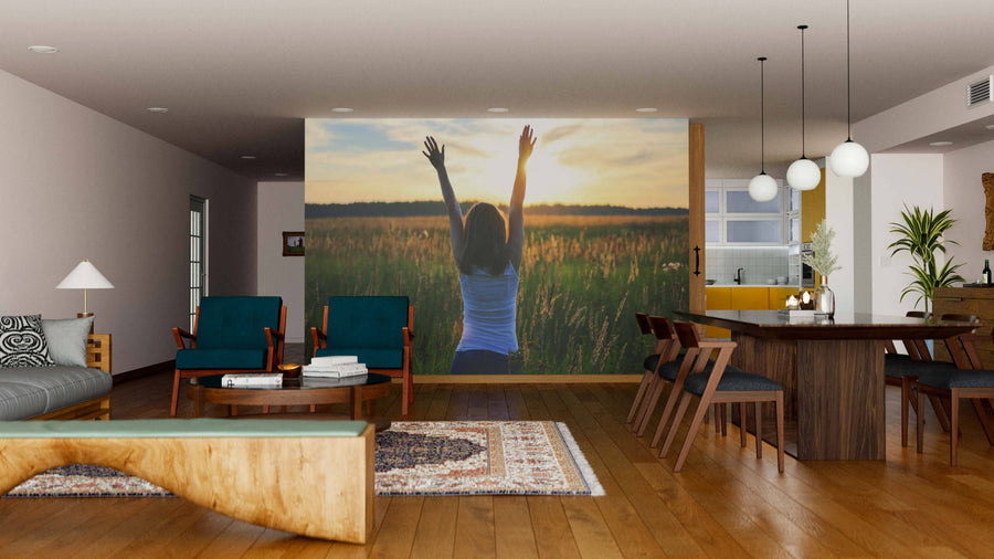 A modern living-dining room with wood floors, retro furniture, a rug, and the Sunrise Gratitude Wall Mural—depicting a woman with raised arms in a sunlit field—creates a cozy open space, highlighted by pendant lights over the dining table.
