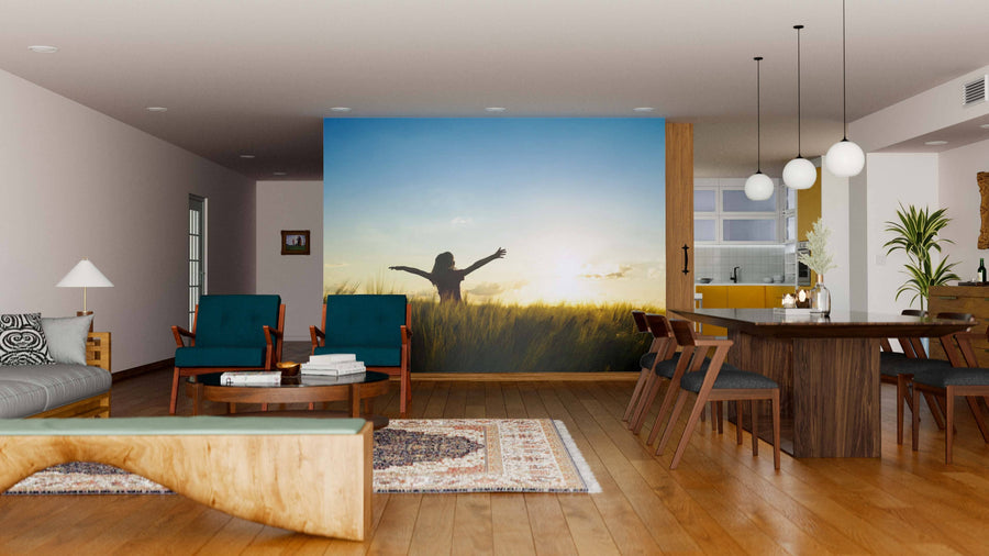 A modern living and dining room with wooden floors, mid-century furniture, teal chairs, a wooden table, pendant lights, and an inviting atmosphere completed by the Sunset View Wall Mural featuring a figure in a sunlit field.
