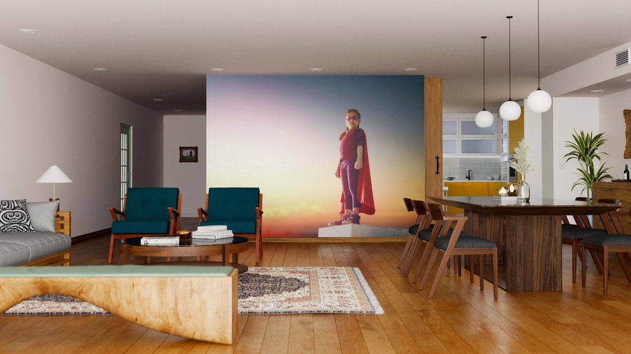 A modern living and dining room with wooden floors, teal chairs, a sofa, and a Superhero Wall Mural showing a child in a cape and mask at sunset. The dining area has a wooden table and globe pendant lights.