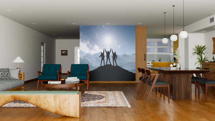 Modern living-dining area with wood floors, teal armchairs, dining table, and kitchen in the background. The "Team at the Peak Wall Mural" displays three figures silhouetted on a mountain against a sunny, snowy peak backdrop.