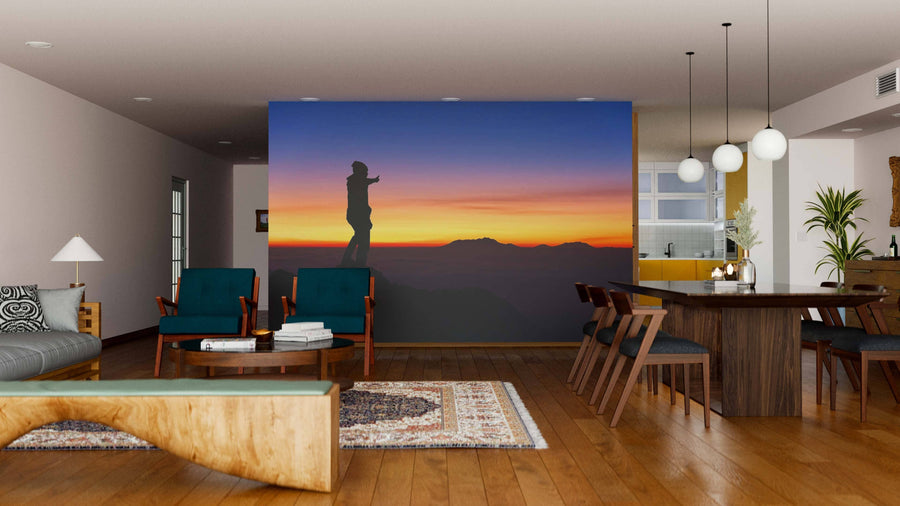 A modern living and dining room with wooden floors, chic furniture, and a striking accent wall featuring the Thumbs Up Wall Mural—showing a person silhouetted on a mountain at sunset with vibrant orange, purple, and blue skies.