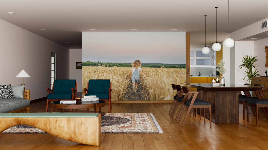 A modern living and dining room with wooden floors, teal armchairs, a large table, and a statement Wheatland Wanderer Wall Mural featuring a child in a wheat field. Warm lighting and minimalist décor complete the cozy, inviting space.