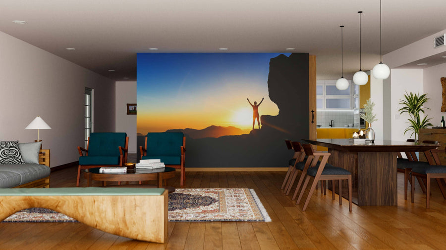 A modern living-dining room with wood floors and mid-century furniture features the Woman Climber Wall Mural, showing a person celebrating on a rocky summit at sunset. Ambient lighting and a rug bring warmth to the inviting space.