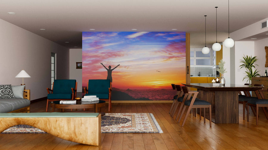 A modern living-dining room features wood furniture, rugs, and the "Woman on Beautiful Sunset Wall Mural," showing a person with raised arms at an ocean sunset with vibrant clouds and a bird, creating a stunning focal point.