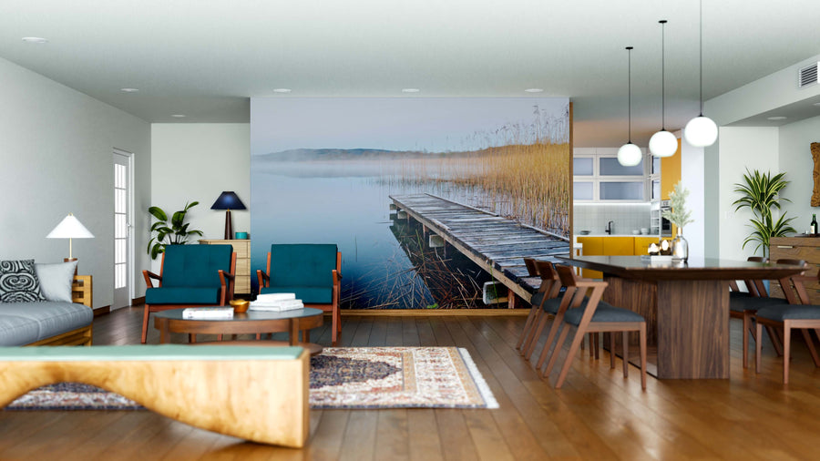 A modern open-plan living and dining area features wood floors, blue armchairs, a wooden coffee table, dark dining set, yellow kitchen island, pendant lights, and an Irish Lake Wall Mural of a lakeside dock as a custom decorative touch.