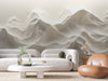 A modern living room includes a white curved sofa, a round coffee table with green plants, and a brown leather chair by a wooden side table decorated with dried flowers. The serene atmosphere is enhanced by the Ivory Dunes Wall Mural featuring abstract wavy mountains in muted colors.