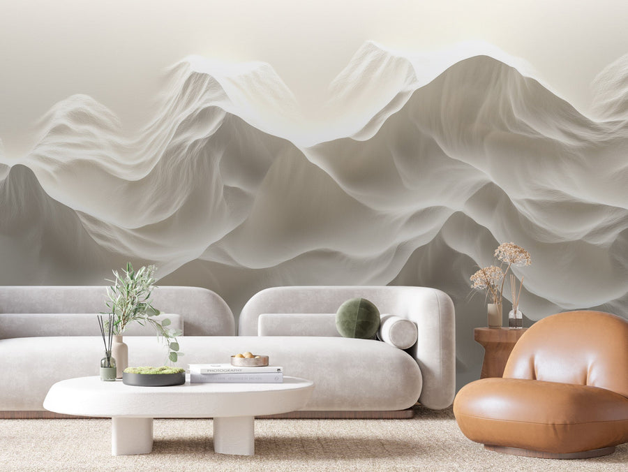 A modern living room includes a white curved sofa, a round coffee table with green plants, and a brown leather chair by a wooden side table decorated with dried flowers. The serene atmosphere is enhanced by the Ivory Dunes Wall Mural featuring abstract wavy mountains in muted colors.