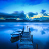 A serene lakeside view at twilight features a wooden dock extending into calm blue waters and a small rowboat, reminiscent of the Jetty and Boat Wall Mural by Deposit Photo. The sky has wispy clouds and distant hills in hues of blue and soft yellow, creating a tranquil scene.