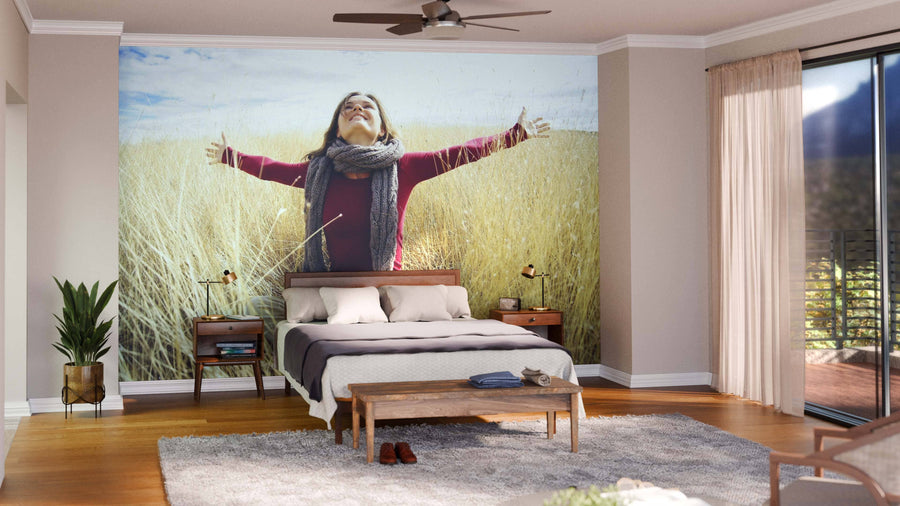 A cozy bedroom features the Joyful Sunshine Wall Mural behind the bed, depicting a joyful woman with arms outstretched in a golden wheat field. Wooden furniture, plants, a ceiling fan, and large windows with sheer curtains complete the space.