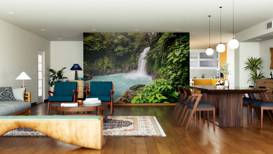 Modern, open-plan living and dining area with wood floors, blue chairs, a tan sofa, and hanging lights. The Jungle of Costa Rica Wall Mural creates a stunning green forest and waterfall focal wall, bringing nature indoors in style.