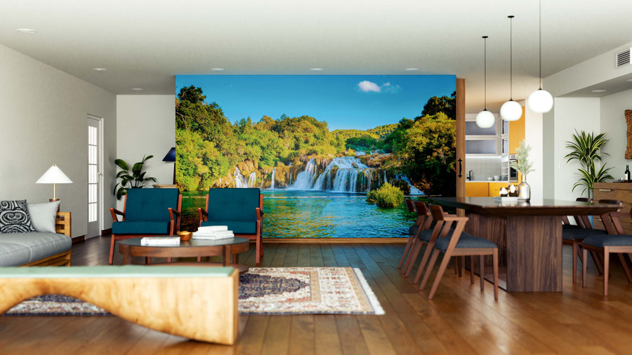 A modern open-concept living and dining room showcases the KRKA Waterfalls Wall Mural, a vibrant, peel-and-stick removable wallpaper that adds lush color to the space, complementing wood floors, mid-century furniture, pendant lights, and a cozy rug.