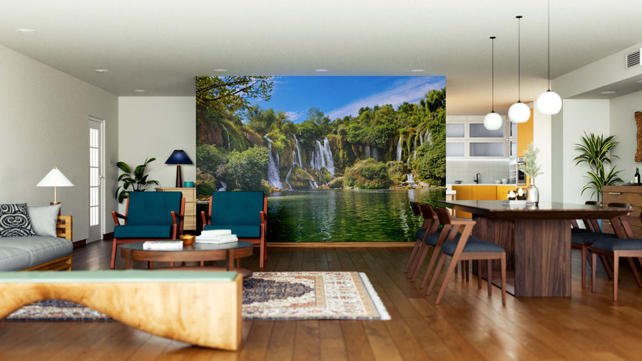 A spacious, modern living-dining room with wood floors and mid-century furniture is brightened by a large Kravice Waterfalls Wall Mural. Teal, wood, and neutral decor complement the waterfall scene as natural light streams in.