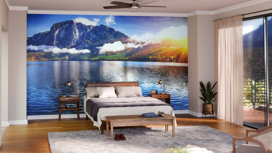 A modern bedroom with a bed, nightstands, and bench features the Lake Altausseer and Totes Gebirge Wall Mural of a lake, mountains, and sunrise. Large windows with sheer curtains, a plant, rug, and ceiling fan complete the cozy decor.