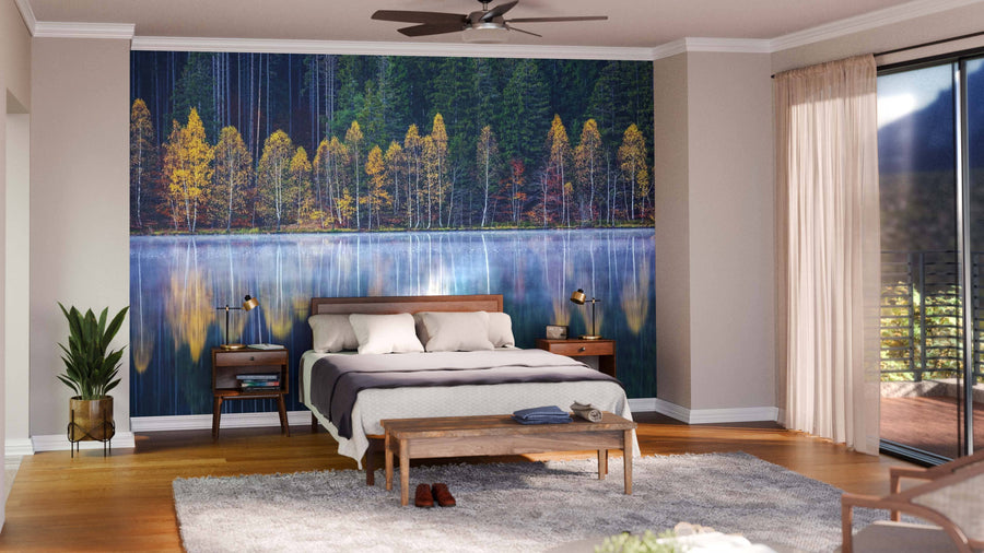 A bedroom features a bed with gray and white bedding, wooden furniture, lamps, and the Lake Reflection Wall Mural behind the bed. Light streams in through sheer-curtained windows, illuminating a plant on the hardwood floor.