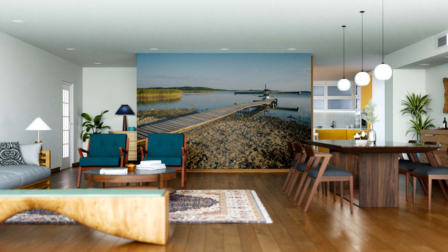Modern open-plan living and dining room with wooden floors, blue and wood furniture, hanging lights, and a large Lakeside Lift-Off Wall Mural featuring a dock over calm water under a blue sky. Kitchen with yellow accents in the background.