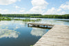 A wooden dock extends over a calm lake, mirroring fluffy clouds, green reeds, and lily pads. This serene landscape resembles the Lakeside Pier Wall Mural, with a dense forest lining the distant shore against a bright blue sky.