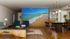 A modern open-concept living and dining area with wooden floors, stylish furniture, and the Australian Beach Scene Wall Mural showcasing turquoise water and golden sand. Spherical pendant lights hang above the dining table.