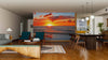 A modern living-dining room with wooden floors, teal chairs, a dining table, and pendant lights features the Beautiful Sunset Over Baltic Sea Wall Mural as a vibrant focal point, creating an open and inviting atmosphere.