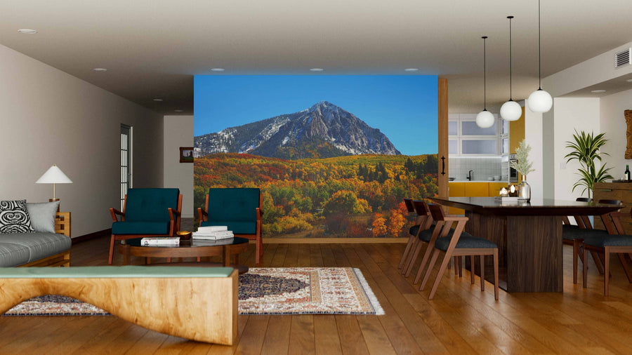 Modern living and dining room with wood flooring and mid-century furniture. The Beckwith Mountain Wall Mural adds vibrant color, covering one wall with a scenic mountain and autumn forest. Pendant lights hang above a dark dining table, chairs, and a rug.