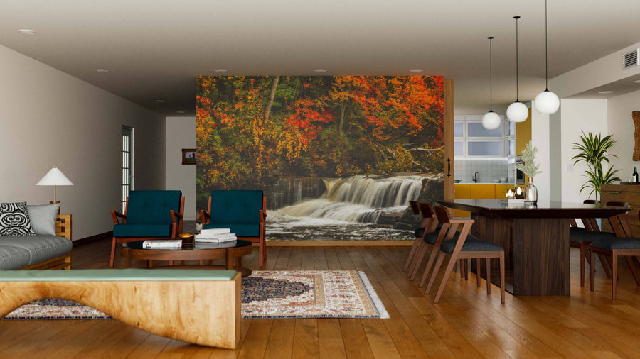 A modern open-plan living and dining room with wood floors, green chairs, a large dining table, and the Berea Falls In Autumn Wall Mural showcasing a waterfall amid vivid autumn trees. Warm hanging globe lights complete the inviting atmosphere.