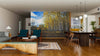 A modern living and dining area with wood floors, teal armchairs, a wooden table, pendant lights, and a sofa features the Birch Trees Blue Sky Fall Wall Mural on one wall, bringing a vibrant autumn scene to the warmly lit space.