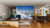 A modern living and dining room with wooden floors and mid-century furniture features the Blue Sky Winter in the Alps Wall Mural, showcasing snowy mountains and pine trees under a blue sky for added depth and a wintry vibe.