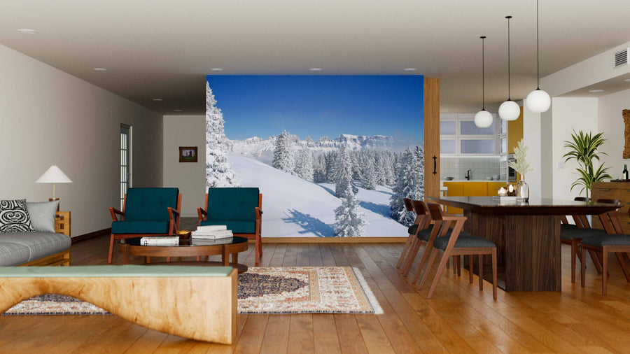 A modern living and dining room with wooden floors and mid-century furniture features the Blue Sky Winter in the Alps Wall Mural, showcasing snowy mountains and pine trees under a blue sky for added depth and a wintry vibe.