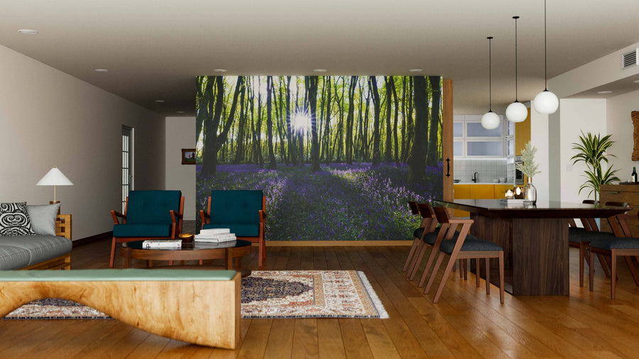 Modern living-dining area with wooden floors, teal armchairs, gray sofa, dining table for six. A Bluebell Wood Forest Wall Mural shows a sunlit forest with green trees and purple flowers. Pendant lights hang above the table; kitchen behind.