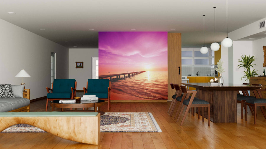 A modern open-plan living and dining room with wood floors, stylish furnishings, and a Boardwalk on Beach Wall Mural featuring a vibrant pink and orange sunset over a pier as a striking accent wall behind the living area.