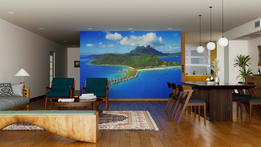 A modern living-dining room with wooden floors and mid-century furniture features a Bora Bora Wall Mural of a tropical island. Teal chairs, a wooden dining table, pendant lights, and rug complete the look; kitchen visible in back.