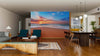 A modern living-dining room with wood floors and teal chairs features the Brilliant Ocean Beach Sunrise Wall Mural, a peel-and-stick design that adds vibrant color to the space. Warm lighting and rustic accents create a cozy, inviting atmosphere.