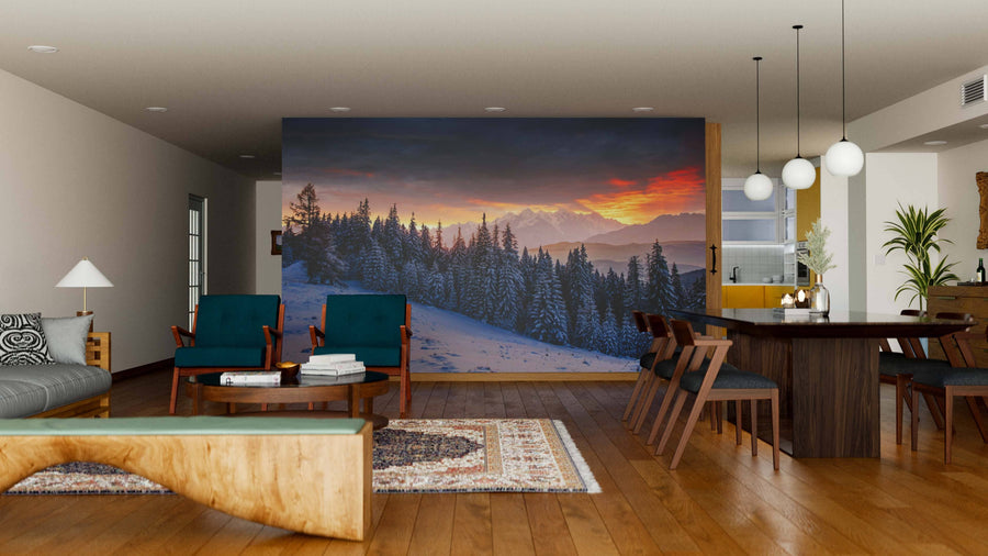 A modern living-dining room with wood floors features teal and wood furniture, a patterned rug, and the "Colorful Sunset Over the Mountain Wall Mural," creating a cozy atmosphere. Pendant lights hang above a dark dining table with six chairs.