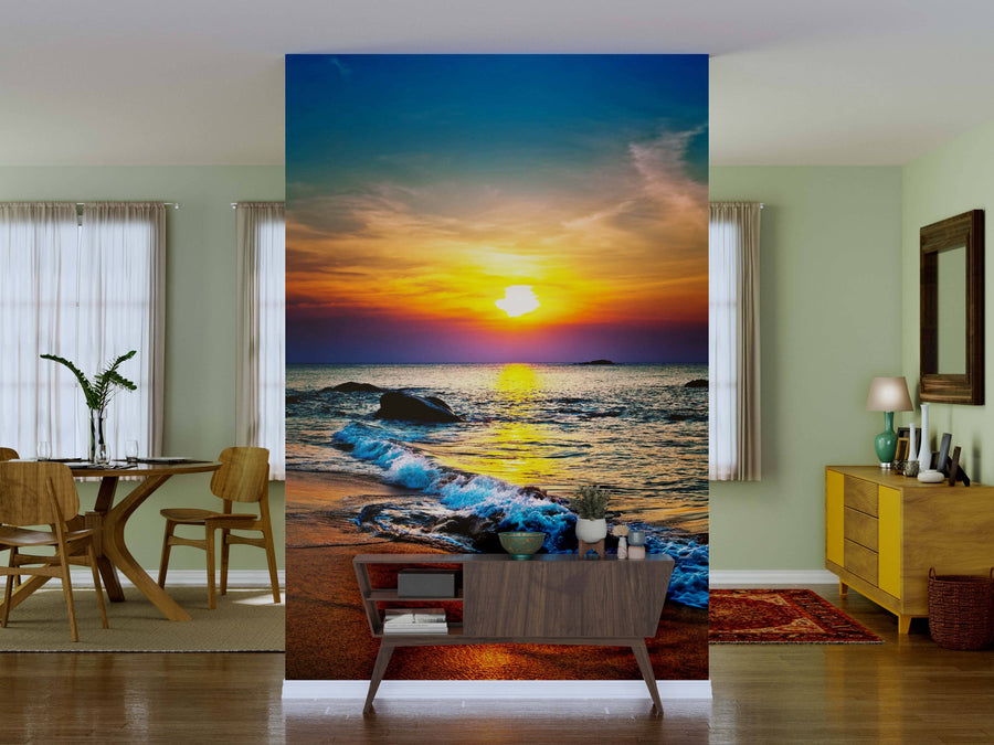 A living room with pale green walls features the Colorful Sunset Over the Sea Wall Mural—a vibrant peel-and-stick beach sunset with waves, rocks, and a colorful sky. Wooden furniture, plants, a red rug, and gentle natural light complete the space.